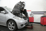 A mechanic in a beanie works under the open hood of a silver car in an auto repair shop.