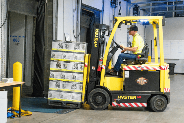 Worker in a high-visibility vest operating a yellow Hyster forklift carrying stacked cases in a warehouse.
