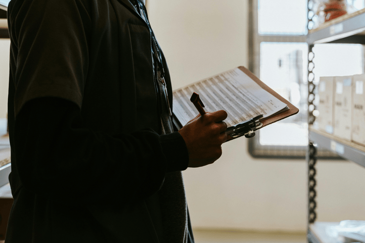 Worker holding a clipboard with a checklist form near storage shelves in a warehouse.